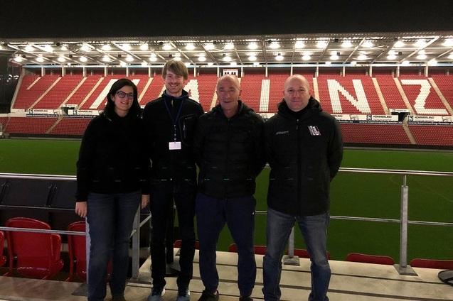 Vier Personen stehen am Rande eines leeren Fu&szlig;ballfeldes in einem Stadion mit beleuchtetem Trib&uuml;nenbereich.