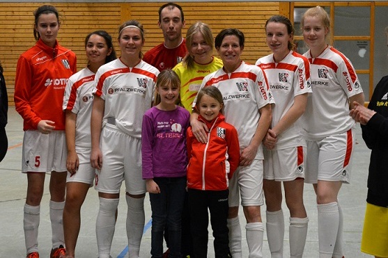 Eine Gruppe von sieben Fu&szlig;ballspielerinnen und zwei M&auml;dchen posiert l&auml;chelnd in Sportkleidung in einer Halle.