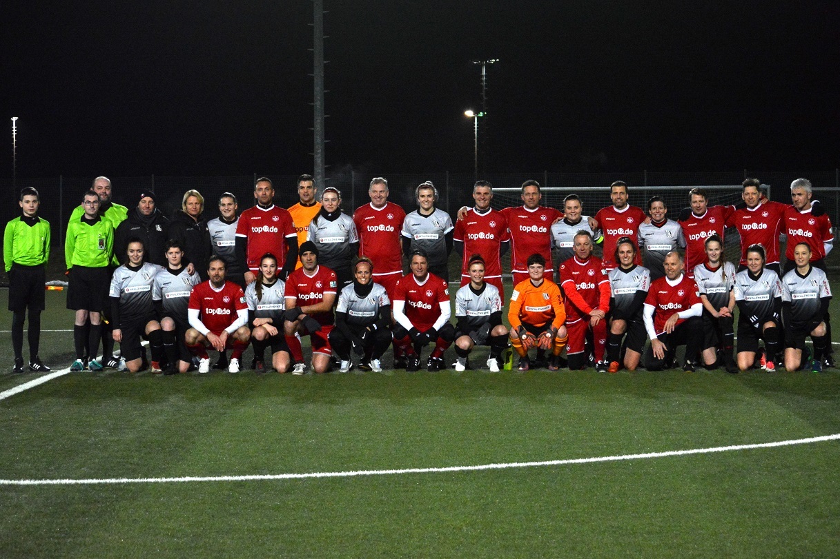 Gruppenfoto von zwei Fu&szlig;ballmannschaften mit Schiedsrichtern auf einem Kunstrasenplatz bei Nacht.