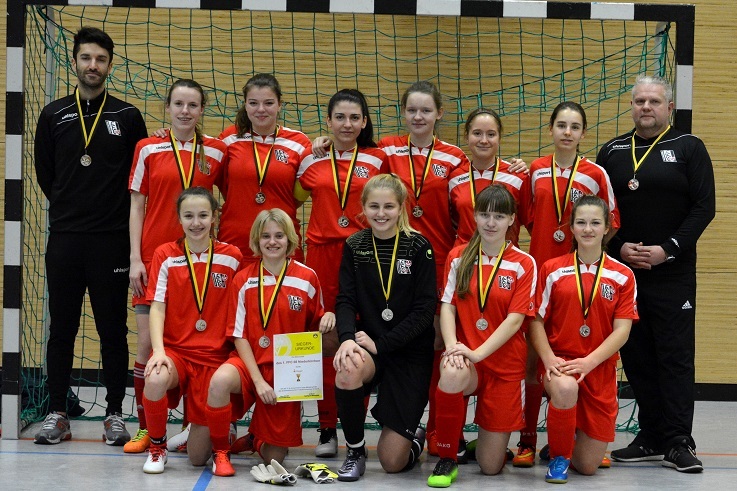 M&auml;dchenfu&szlig;ballmannschaft mit Medaillen und Urkunde, posierend vor einem Handballtor in roten Trikots.