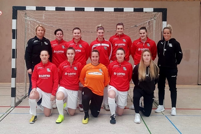 Teamfoto einer Frauenfu&szlig;ballmannschaft in roten Trikots vor einem Tor in einer Sporthalle.