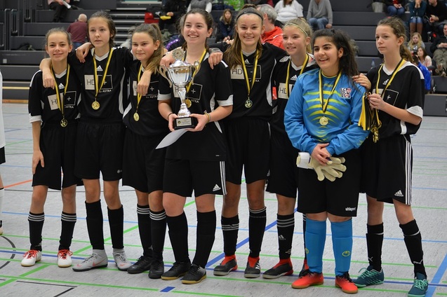 Sieben M&auml;dchen in Fu&szlig;balluniformen stehen lachend zusammen, halten einen Pokal und tragen Medaillen.