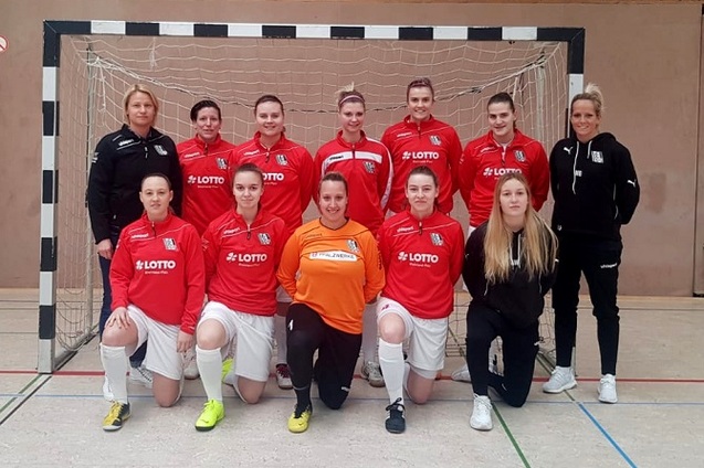 Teamfoto einer Frauenfu&szlig;ballmannschaft in roten Trikots vor einem Tor in einer Sporthalle.