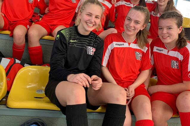 Gruppe von M&auml;dchen in roten Fu&szlig;balltrikots sitzt l&auml;chelnd auf einer Trib&uuml;ne in einer Sporthalle.
