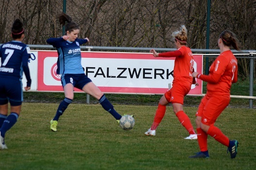 Fussballspiel: Spielerin in blauer Uniform tritt nach einem Ball, w&auml;hrend zwei Gegnerinnen in roten Trikots n&auml;herkommen.