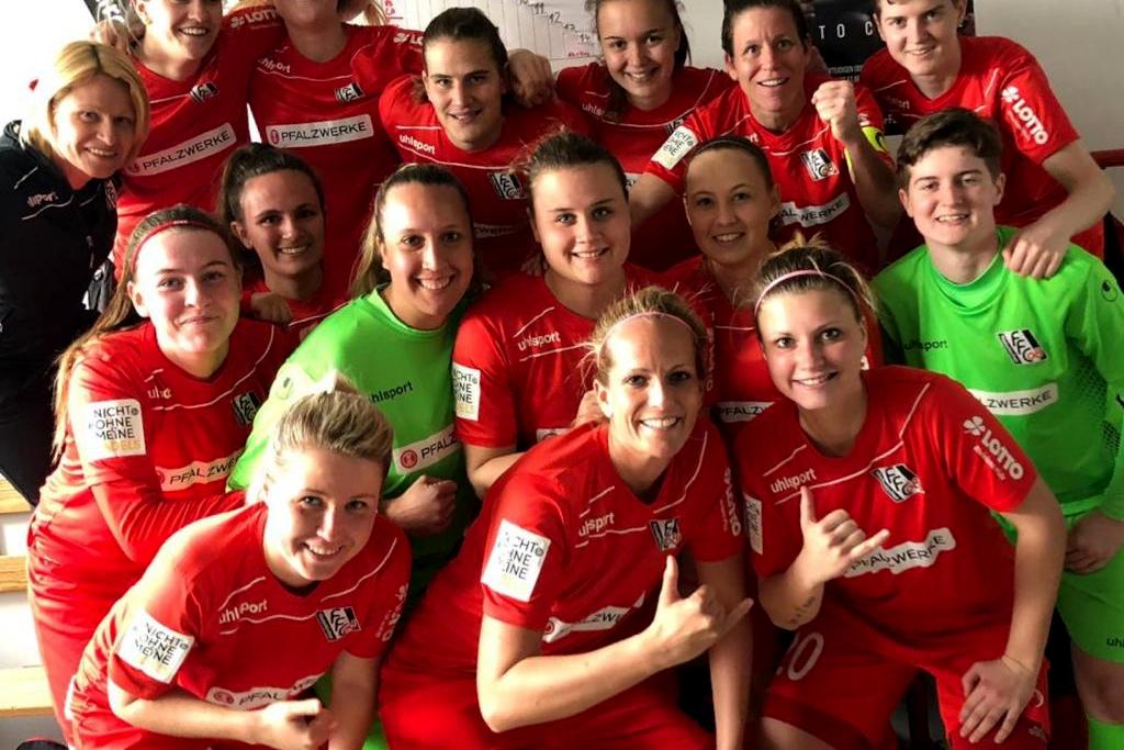 Gruppenfoto von 16 Frauen in roten Fußballtrikots, lächelnd und in zwei Reihen vor einer Wand mit Sportplakaten.