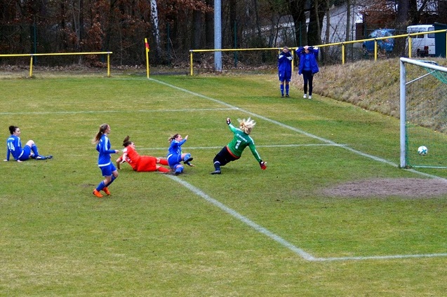 Torfrau in gr&uuml;n springt, um einen Ball zu halten, w&auml;hrend Spielerinnen am Boden liegen. Zuschauer im Hintergrund.
