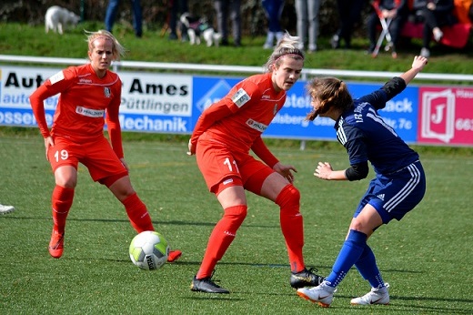 Drei Fu&szlig;ballspielerinnen im Wettkampf auf einem Kunstrasenplatz, eine Spielerin in Blau dribbelt den Ball.