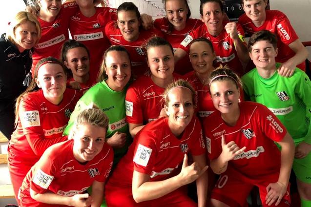 Gruppenfoto von 16 Frauen in roten Fu&szlig;balltrikots, l&auml;chelnd und in zwei Reihen vor einer Wand mit Sportplakaten.
