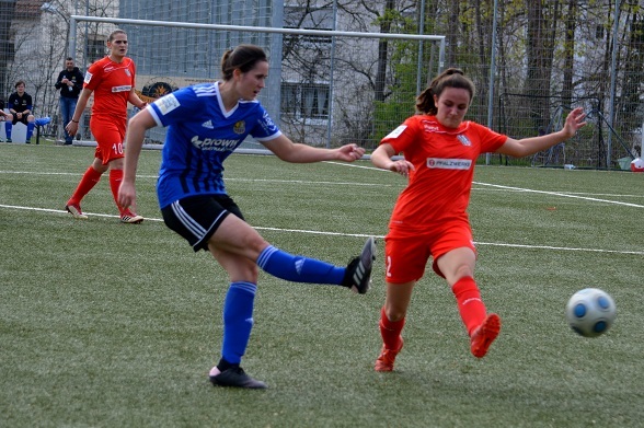 Zwei Fu&szlig;ballspielerinnen im Wettkampf, w&auml;hrend die eine den Ball mit dem Fu&szlig; trifft, der auf dem gr&uuml;nen Spielfeld liegt.