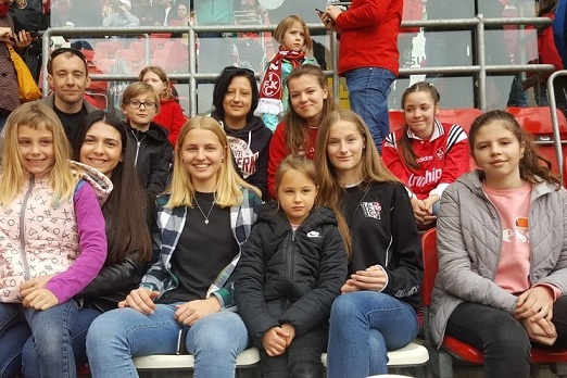 Gruppe von zehn Personen, darunter M&auml;dchen und Frauen, sitzend im Stadion, mit Fanartikeln in Rot und Schwarz.