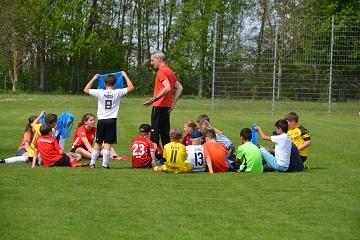 Kinder in Sporttrikots sitzen im Kreis auf einem Fu&szlig;ballplatz, w&auml;hrend ein Trainer ihnen Anweisungen gibt.