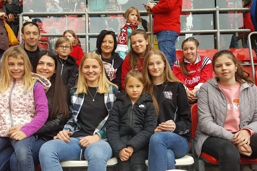 Gruppe von zehn Personen, darunter M&auml;dchen und Frauen, sitzend im Stadion, mit Fanartikeln in Rot und Schwarz.