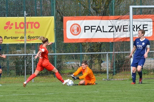 Spielszene im Frauenfu&szlig;ball: Eine Spielerin in Rot schie&szlig;t auf das Tor, w&auml;hrend die Torh&uuml;terin in Orange den Ball abwehrt.