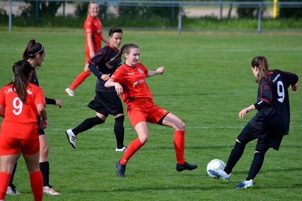 Vier Fu&szlig;ballspielerinnen auf einem gr&uuml;nen Feld, eine in rot dribbelt den Ball, w&auml;hrend eine schwarze Spielerin versucht, ihn zu stoppen.