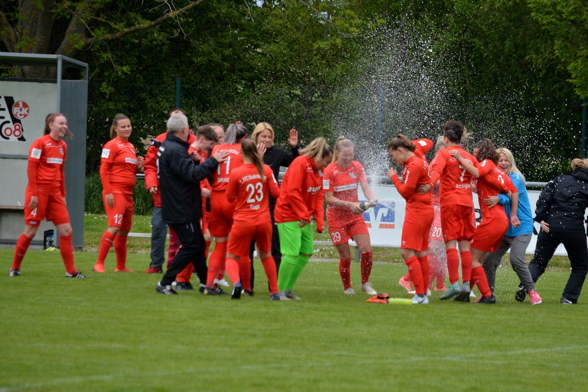 Freuen sich jubelnde Frauenmannschaft in roten Trikots, w&auml;hrend Wasser spritzt, auf einem Fu&szlig;ballplatz.