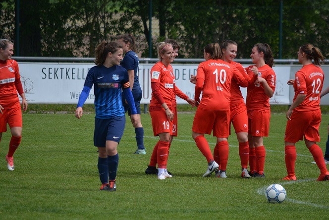 Frauenfu&szlig;ballmannschaft in roten Trikots feiert ein Tor, w&auml;hrend eine Spielerin in blauen Trikots im Hintergrund steht.