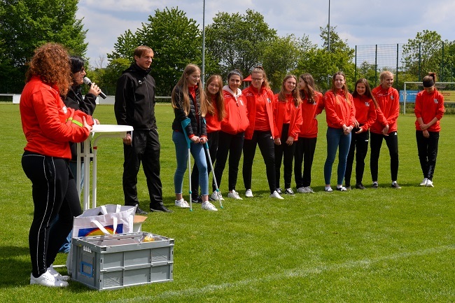Gruppenszene auf einem Sportplatz mit l&auml;chelnden Jugendlichen in roten Jacken und Rednern am Tisch.