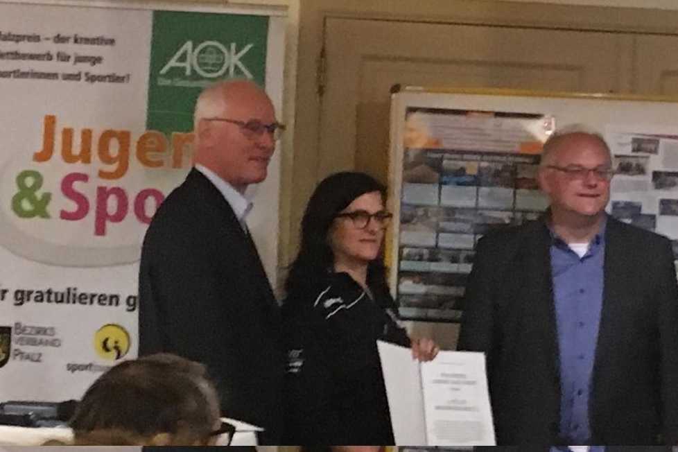 Drei Personen stehen vor einer Wand mit einem Plakat zu &bdquo;Jugend & Sport&ldquo; und halten eine Urkunde in der Mitte.