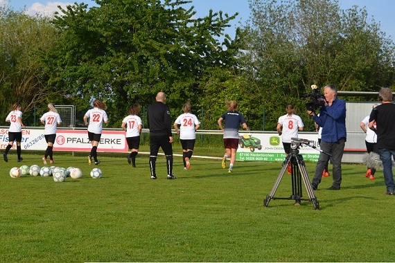 Fu&szlig;ballspieler in Trikots laufen auf einem Platz, w&auml;hrend ein Kamerateam sie filmt und B&auml;lle im Vordergrund liegen.