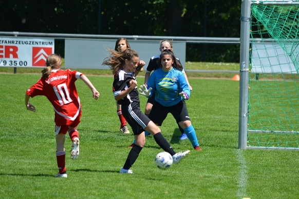 M&auml;dchenfu&szlig;ballspiel: Eine Spielerin in Schwarz schie&szlig;t auf das Tor, w&auml;hrend die Torh&uuml;terin in Blau bereit ist.