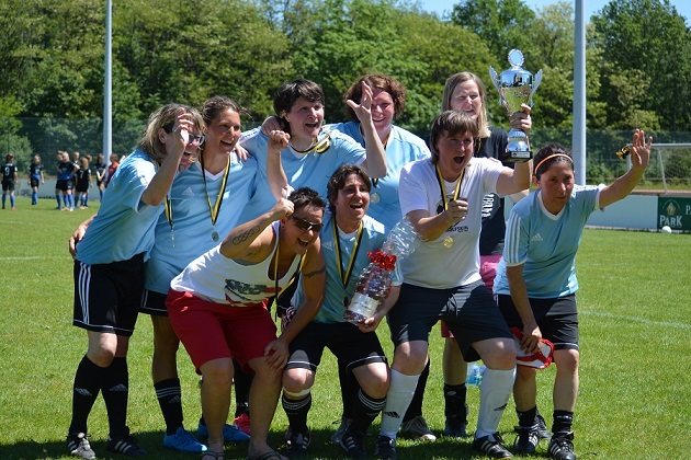 Gruppe von Frauen mit Medaillen und Pokal, jubelnd nach einem Fu&szlig;ballspiel auf einem Rasenplatz.