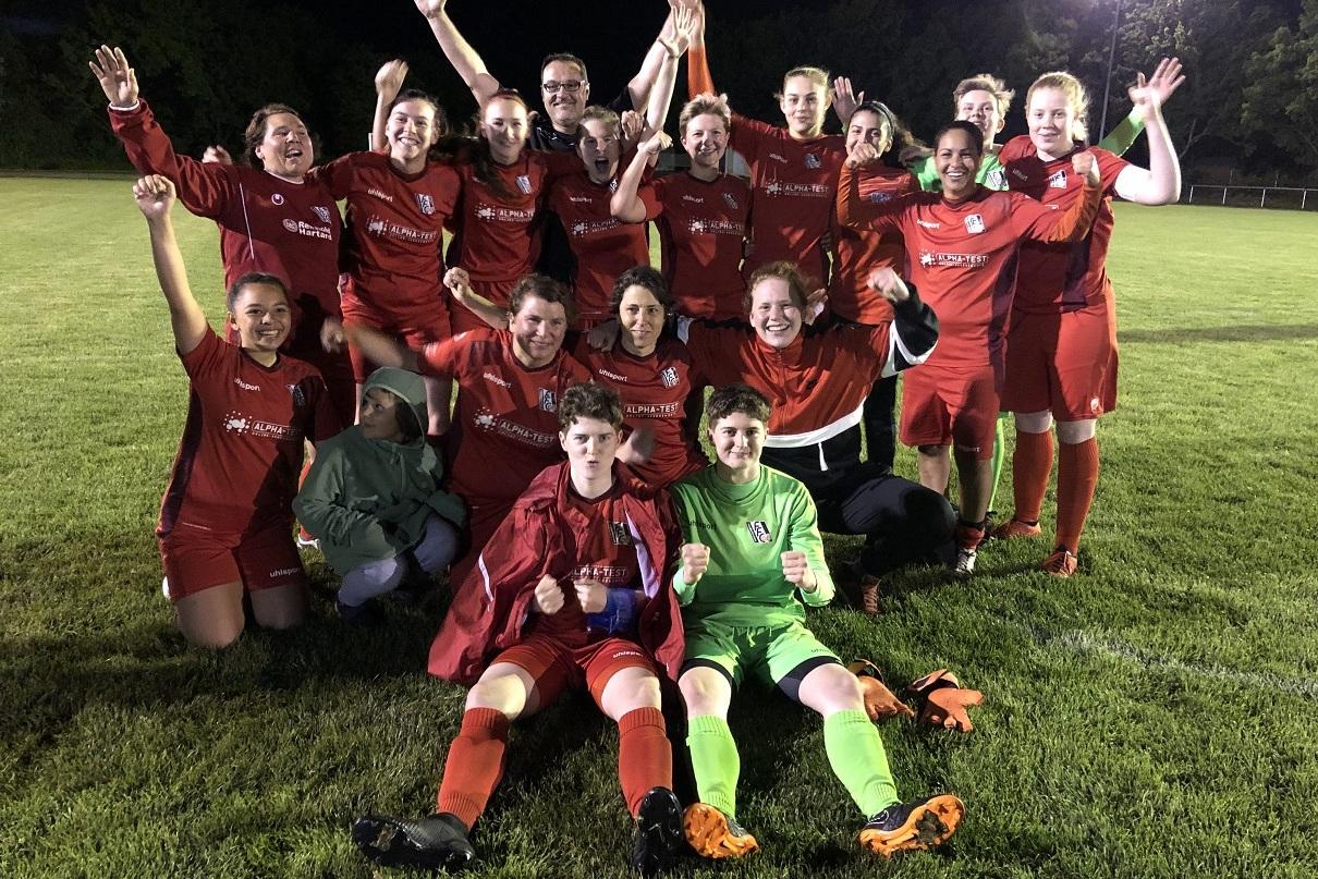 Gruppenspielerinnen in roten Trikots posieren fröhlich nach einem Fußballspiel auf einem Rasenfeld unter Flutlicht.