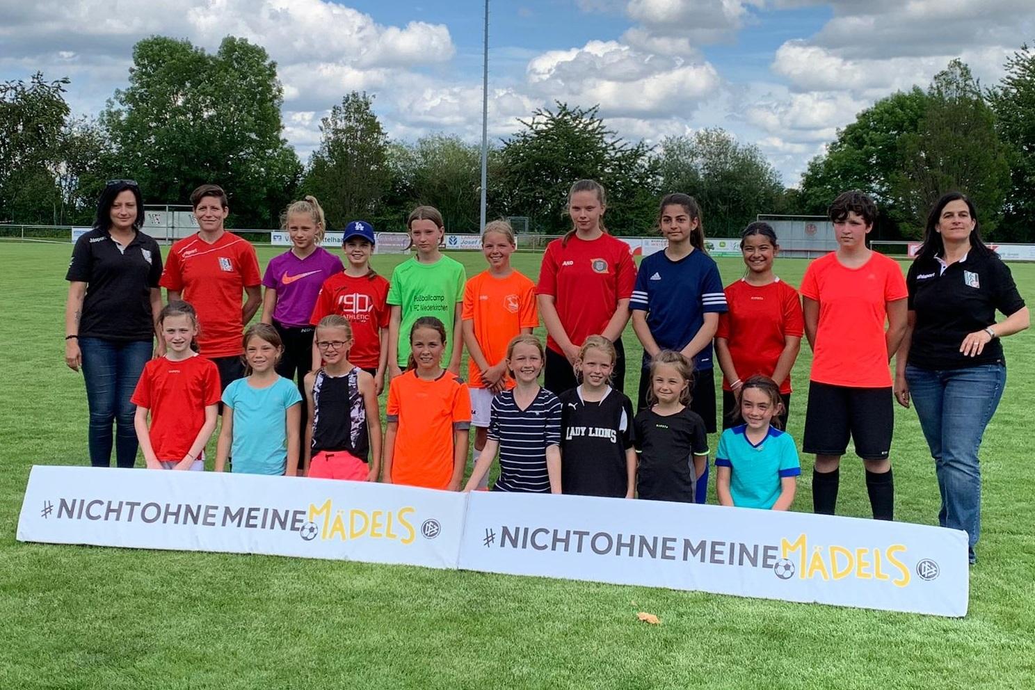 Gruppe von Mädchen in Sportkleidung steht auf einem Fußballfeld vor einem Banner mit der Aufschrift „Nicht ohne meine Mädels“.