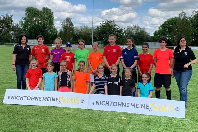 Gruppe von M&auml;dchen in Sportkleidung steht auf einem Fu&szlig;ballfeld vor einem Banner mit der Aufschrift &bdquo;Nicht ohne meine M&auml;dels&ldquo;.