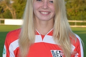 L&auml;chelnde junge Frau in einem roten Fu&szlig;balltrikot steht vor einem Sportplatz mit B&auml;umen im Hintergrund.