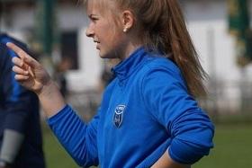 Mädchen mit blauer Sportjacke zeigt mit dem Finger und steht auf einem Fußballfeld.