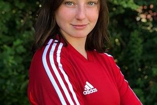 Portr&auml;t eines M&auml;dchens in rotem Fu&szlig;balltrikot mit wei&szlig;en Streifen, verschr&auml;nkten Armen vor gr&uuml;nem Hintergrund.