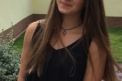 Junge Frau mit langen, braunen Haaren steht l&auml;chelnd vor einem gr&uuml;nen Garten und tr&auml;gt ein schwarzes Top und wei&szlig;e Shorts.