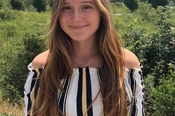 Junge Frau mit langen, braunen Haaren steht mit l&auml;chelndem Gesicht vor gr&uuml;ner Landschaft an einem sonnigen Tag.