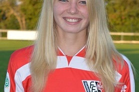 L&auml;chelnde junge Frau in einem roten Fu&szlig;balltrikot steht vor einem Sportplatz mit B&auml;umen im Hintergrund.