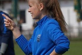 M&auml;dchen mit blauer Sportjacke zeigt mit dem Finger und steht auf einem Fu&szlig;ballfeld.