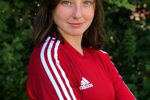 Portr&auml;t eines M&auml;dchens in rotem Fu&szlig;balltrikot mit wei&szlig;en Streifen, verschr&auml;nkten Armen vor gr&uuml;nem Hintergrund.