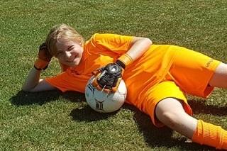 Junger Torwart in orangeem Trikot, liegend auf dem Rasen mit einem Fu&szlig;ball und einer Torwarthandschuh.