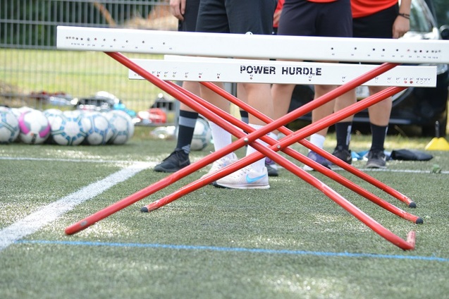 Nahaufnahme einer roten H&uuml;rde mit dem Schriftzug "POWER HURDLE" vor Sportlern auf einem Fu&szlig;ballplatz.