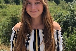 Junge Frau mit langen, braunen Haaren steht mit l&auml;chelndem Gesicht vor gr&uuml;ner Landschaft an einem sonnigen Tag.