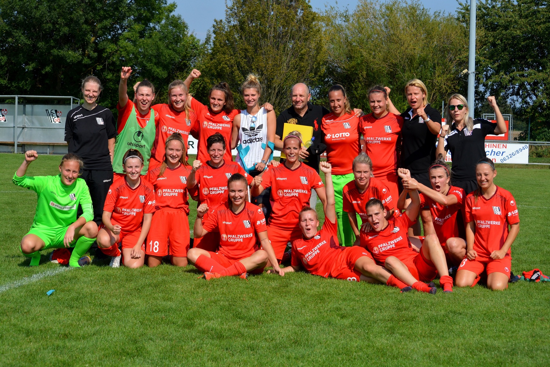 Gruppierung von Fu&szlig;ballspielerinnen in roten Trikots, l&auml;chelnd und jubelnd, auf einem gr&uuml;nen Fu&szlig;ballfeld.
