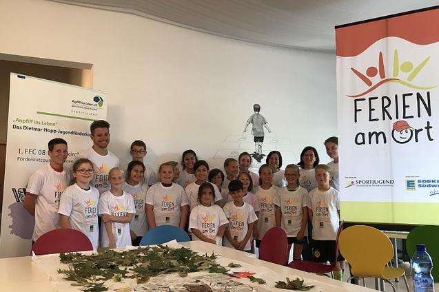 Gruppe von Kindern in T-Shirts mit dem Schriftzug &bdquo;FERIEN am Ort&ldquo; posiert vor einem Banner und einem Tisch mit Bl&auml;ttern.