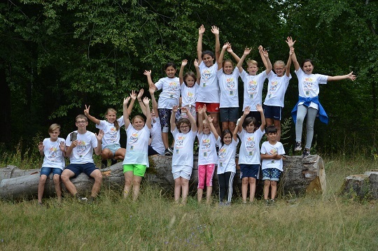 Gruppe von Kindern in wei&szlig;en T-Shirts, die fr&ouml;hlich auf Baumst&auml;mmen im Freien posieren.