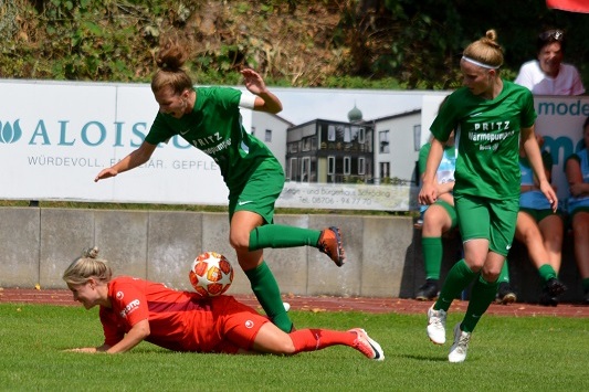 Zwei Spielerinnen in gr&uuml;nen Trikots k&auml;mpfen um den Ball, w&auml;hrend eine Spielerin in rotem Trikot am Boden liegt.