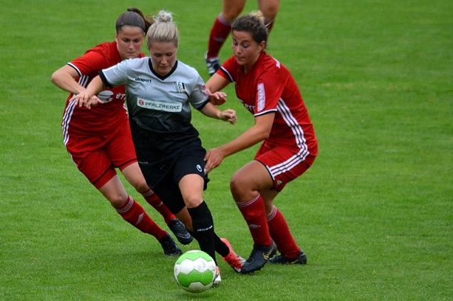 Zwei Spielerinnen in roten Trikots versuchen, eine gegnerische Spielerin in Schwarz am Ball zu hindern.