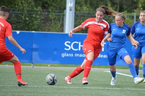 Spielerinnen im Wettkampf: eine mit roten Trikots dribbelt den Ball, w&auml;hrend zwei in blauen Trikots verfolgen.