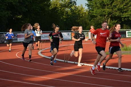 Gruppe von M&auml;dchen joggt auf einer roten Tartanbahn unter strahlendem Sonnenlicht.