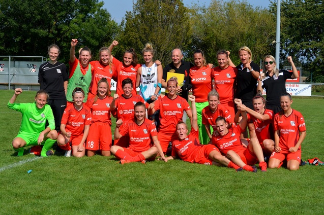 Gruppierung von Fu&szlig;ballspielerinnen in roten Trikots, l&auml;chelnd und jubelnd, auf einem gr&uuml;nen Fu&szlig;ballfeld.