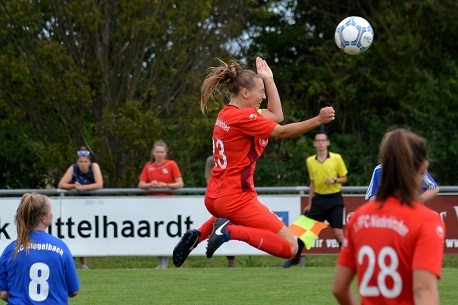 Spielerin in rotem Trikot springt hoch, um einen Ball w&auml;hrend eines Fu&szlig;ballspiels zu k&ouml;pfen, im Hintergrund Zuschauer und Schiedsrichter.