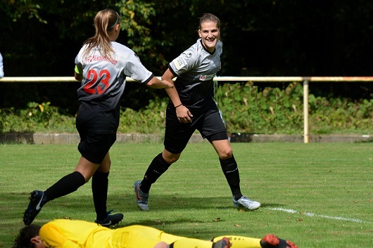 Zwei Fu&szlig;ballspielerinnen feiern einen Torerfolg, w&auml;hrend ein Spieler am Boden liegt. Gr&uuml;ne Wiese im Hintergrund.